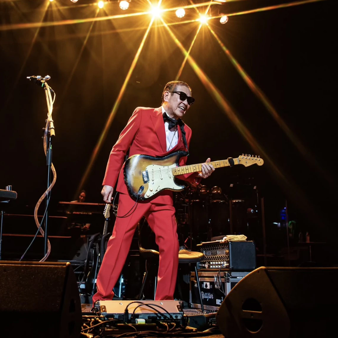 Takanaka in a red suit playing a sunburst electric guitar under stage lights