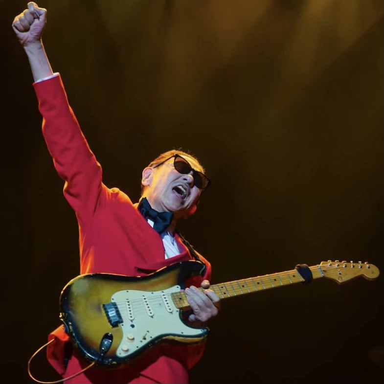 Takanaka in a red suit raising a fist while playing a sunburst electric guitar