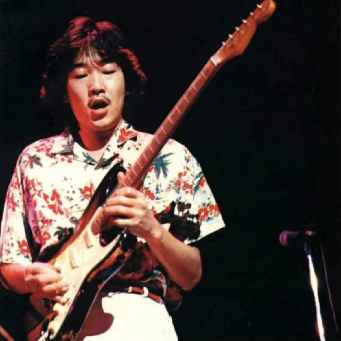Takanaka in a floral shirt playing an electric guitar under red stage lighting